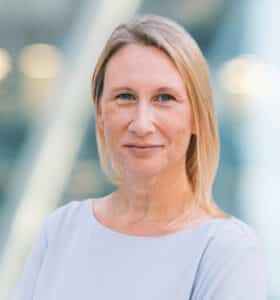 A woman with straight blonde hair, wearing a light grey top, stands in front of a softly blurred background with blue and yellow tones. She is facing the camera and smiling slightly.