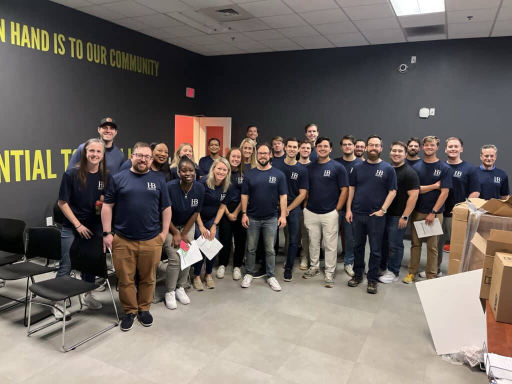 A group of people wearing matching shirts stands in a room with black walls and motivational text. Some hold papers, while boxes and chairs are nearby. They smile at the camera, appearing to be part of a team event or gathering.