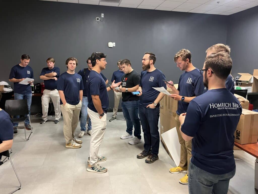 A group of people in matching navy blue shirts with Homrich Berg on the back are gathered in a room. Some are standing and having a conversation, while others are looking at papers. The room has a gray wall and a light-colored floor.