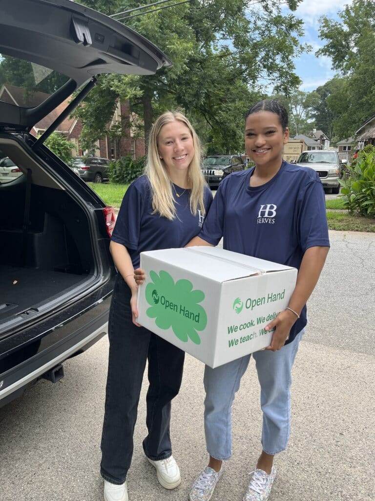 Two women smiling and holding a white box with Open Hand printed on it. They stand next to an open car trunk on a residential street. One woman wears a dark blue shirt, and the other is in light blue jeans. Trees and houses are in the background.