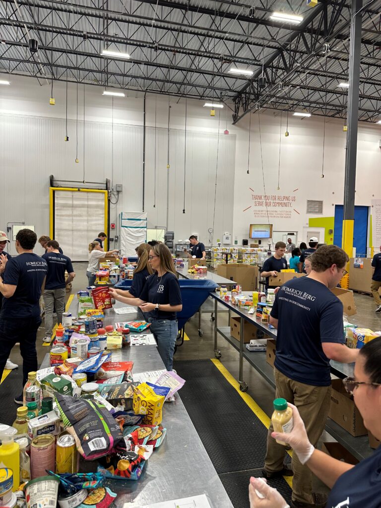 Volunteers in a warehouse sort and organize food items on tables. They are surrounded by boxes filled with canned goods, packaged foods, and beverages. Everyone is wearing matching shirts and focusing on their tasks in the spacious industrial setting.