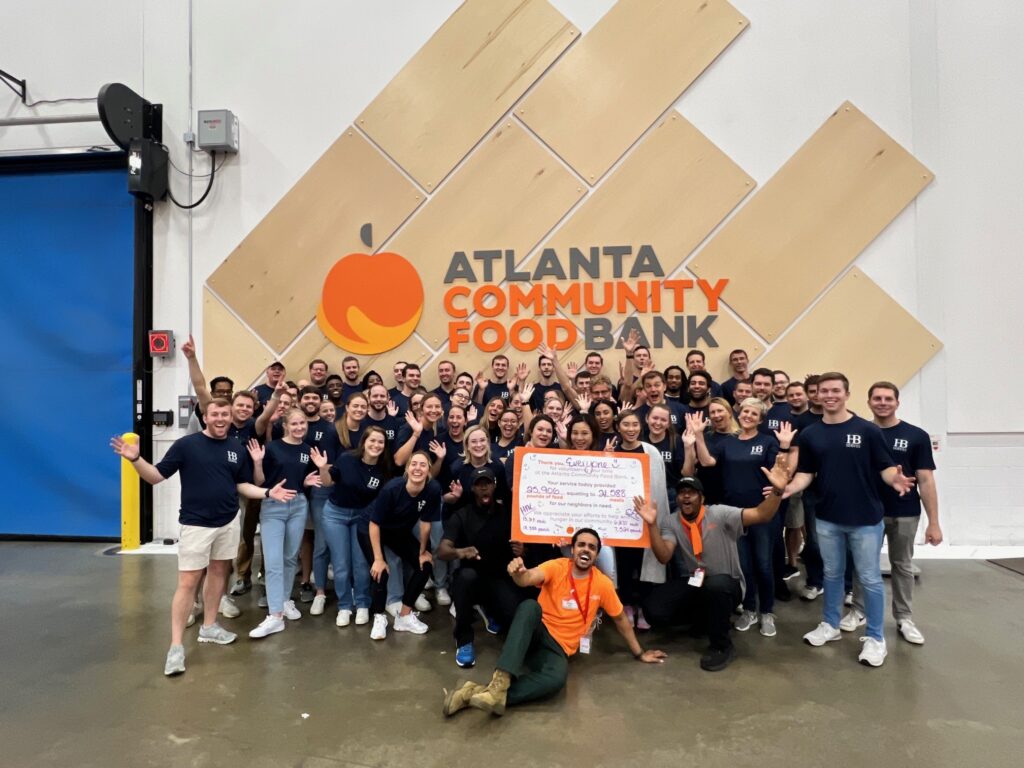 A group of people wearing matching shirts pose enthusiastically in front of an Atlanta Community Food Bank sign. One person kneels in front holding a large check.