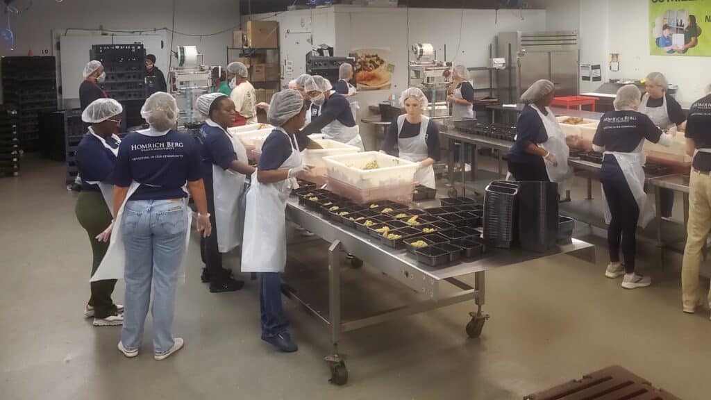 A group of people wearing hair nets and aprons work in a commercial kitchen, assembling meals in black containers. Tables are lined with trays of food, and large kitchen equipment is visible in the background.