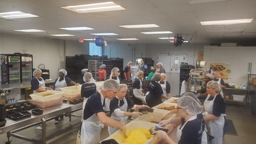 A group of people wearing hairnets and aprons prepare food in a commercial kitchen. They are gathered around large containers, scooping and packaging meals. The room is equipped with industrial kitchen equipment and stainless steel counters.