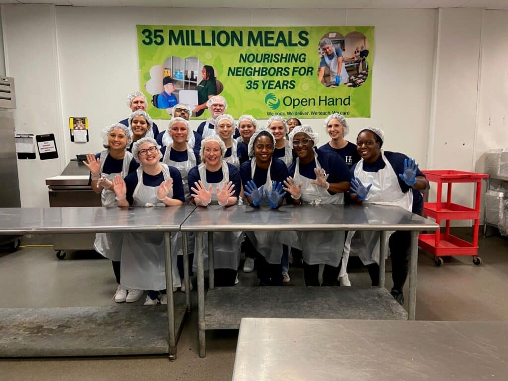 A group of people wearing aprons and hairnets, smiling and posing with gloved hands raised in a kitchen setting. Behind them is a banner celebrating 35 Million Meals from Open Hand, highlighting 35 years of service.