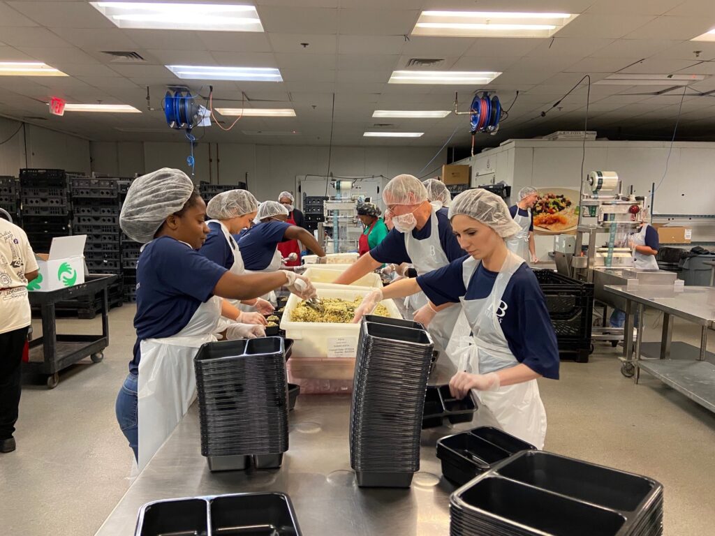 A group of people wearing hairnets, aprons, and gloves work together in a large kitchen. They are preparing food and filling plastic containers. Stacks of empty containers are on the stainless steel table in front of them.