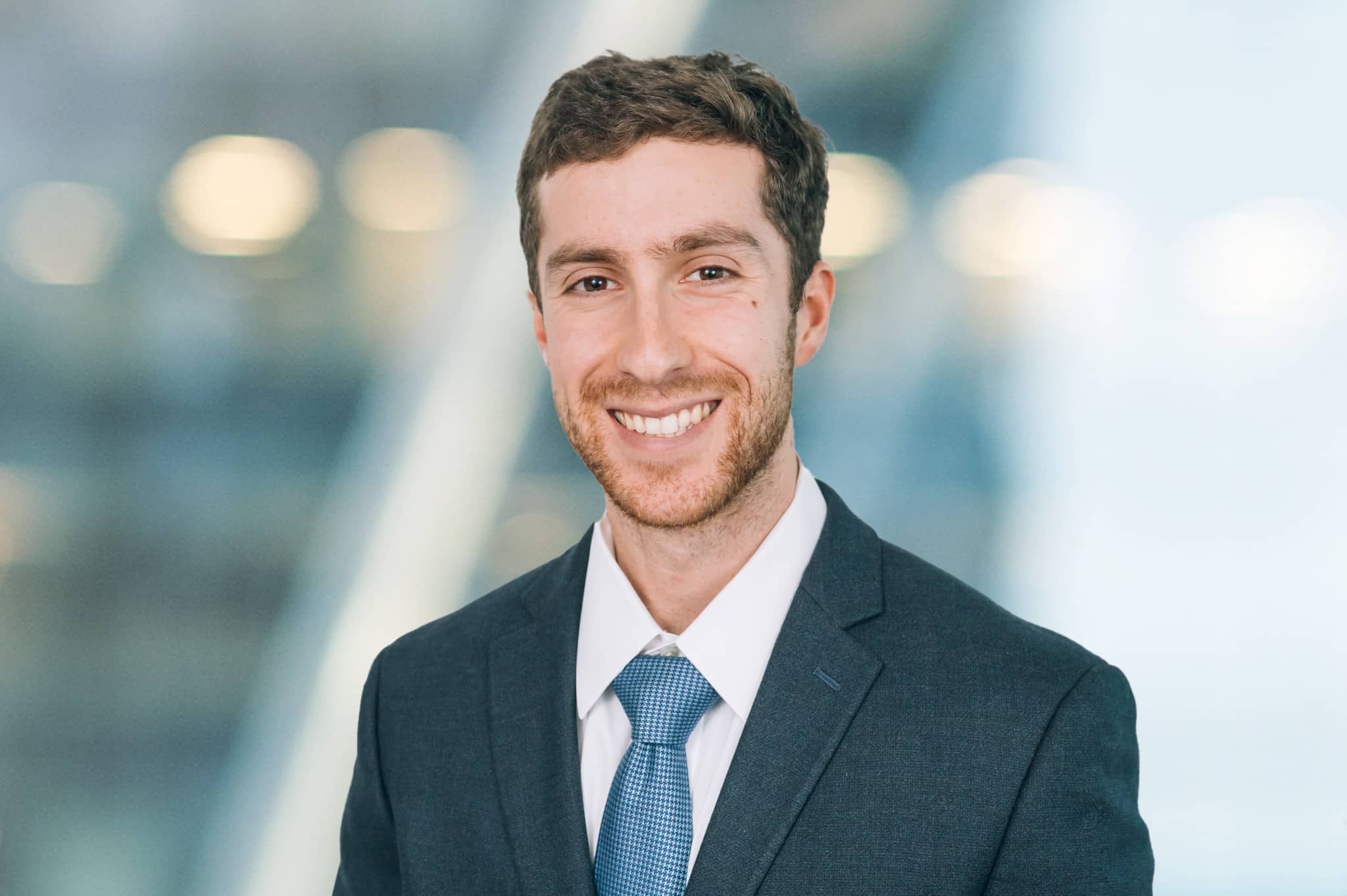 A smiling man in a suit and tie stands in front of a blurred background with lights. He has short brown hair and a neatly trimmed beard.