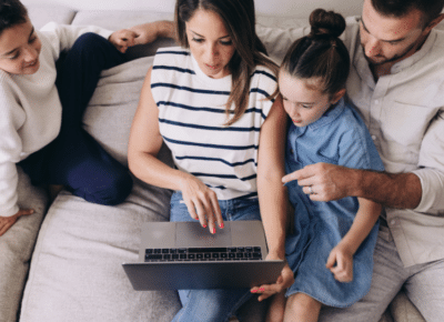 A family of four sits on a couch, gathered around a laptop. The mother points at the screen, while the father and two children, a boy and a girl, look on with interest. They appear engaged and focused on the laptop.