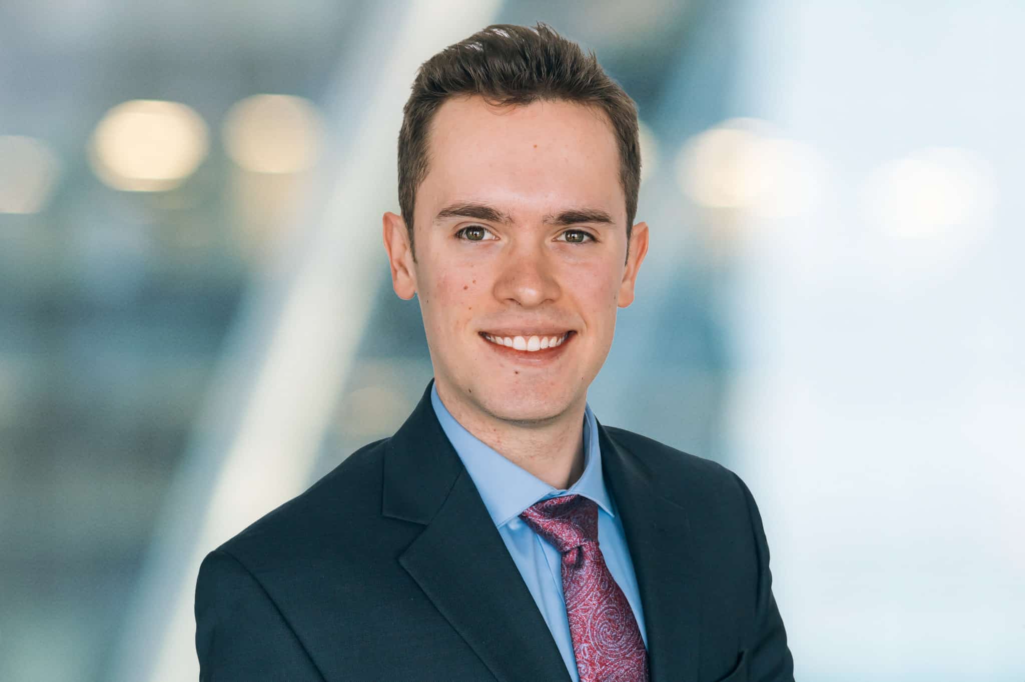 A young man wearing a dark suit, blue shirt, and patterned tie is smiling with a blurred background of bright lights.