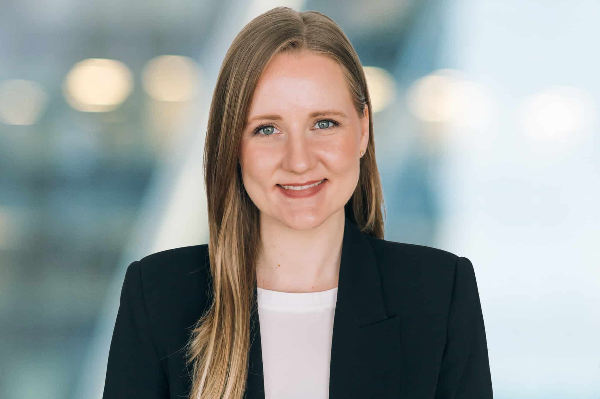 A person with long, straight hair and a slight smile is wearing a black blazer over a white shirt. The background is softly blurred with abstract, circular lights.