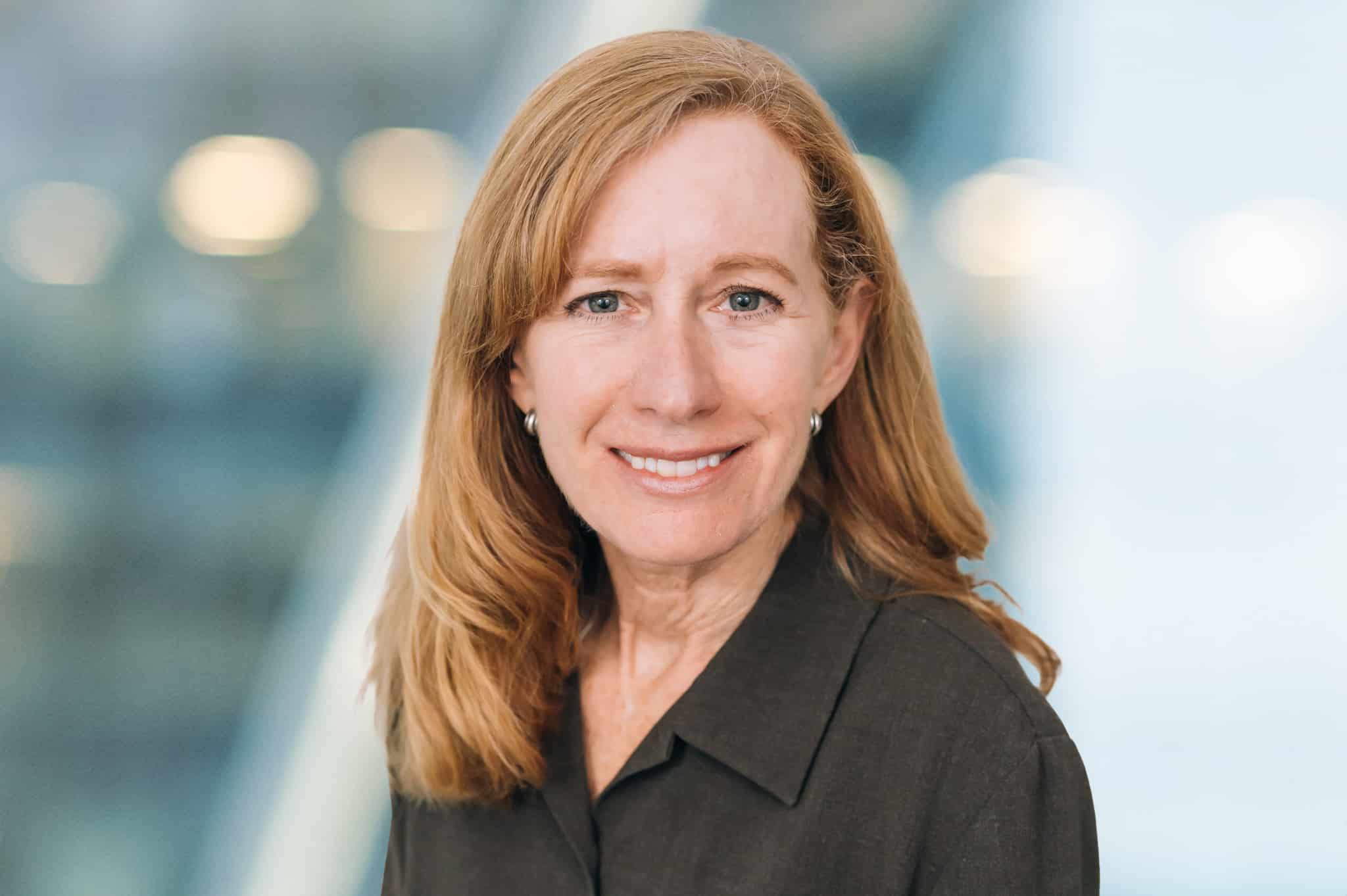 A person with long auburn hair and wearing a dark blouse is smiling at the camera. The background is blurred with soft lights.