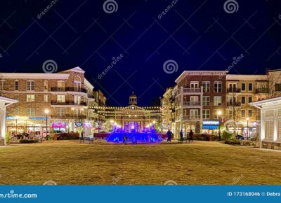 A lively town square at night features a blue-lit fountain, surrounded by shops, restaurants, and string lights between buildings. People are walking and the scene has a warm, festive atmosphere.
