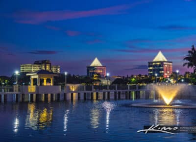 A twilight view of downtown West Palm Beach, Florida, featuring a fountain in a calm lake, illuminated pavilion, and modern buildings with lights reflecting on the water.