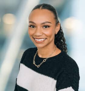 A woman with curly hair tied back, wearing a black and white striped sweater and a gold chain necklace, smiles at the camera. The background is blurred with blue and white tones.
