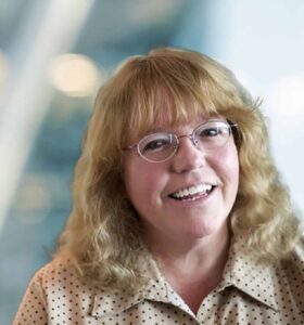 A smiling woman with glasses and wavy blonde hair, wearing a beige blouse with black polka dots, sits in front of a blurred background with soft lights.