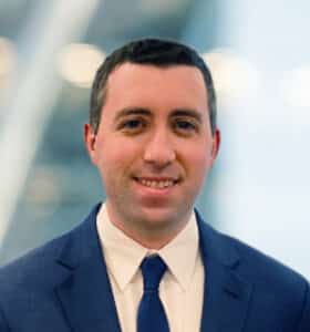 A man with short dark hair, wearing a navy blue suit, white shirt, and dark tie, smiles at the camera against a blurred office background with bright lights.