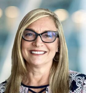 A smiling woman with long blonde hair and glasses, wearing a patterned top, standing in front of a blurred indoor background with soft lighting.