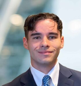 A young man in a suit and tie smiles at the camera. The background is blurred, featuring soft lights and abstract shapes, suggesting an indoor office or professional setting.