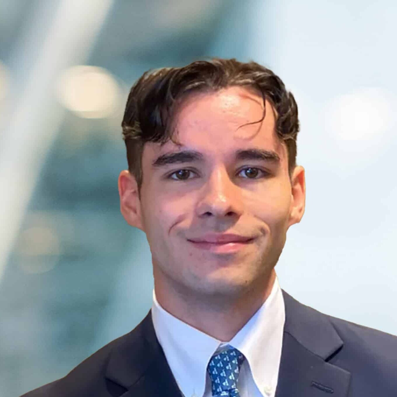 A young man in a suit and tie smiles at the camera. The background is blurred, featuring soft lights and abstract shapes, suggesting an indoor office or professional setting.