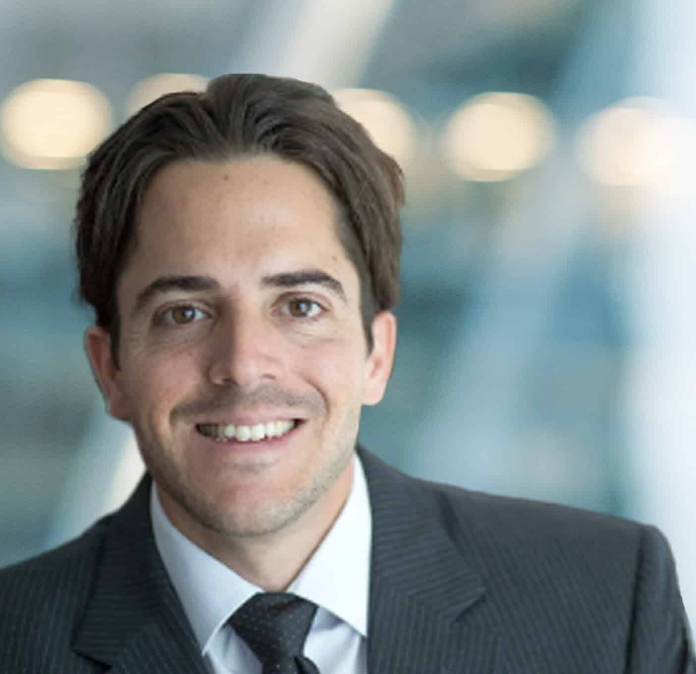 A man with dark hair wearing a dark pinstripe suit, white shirt, and dark tie, smiling at the camera, with a blurred light blue and white background.