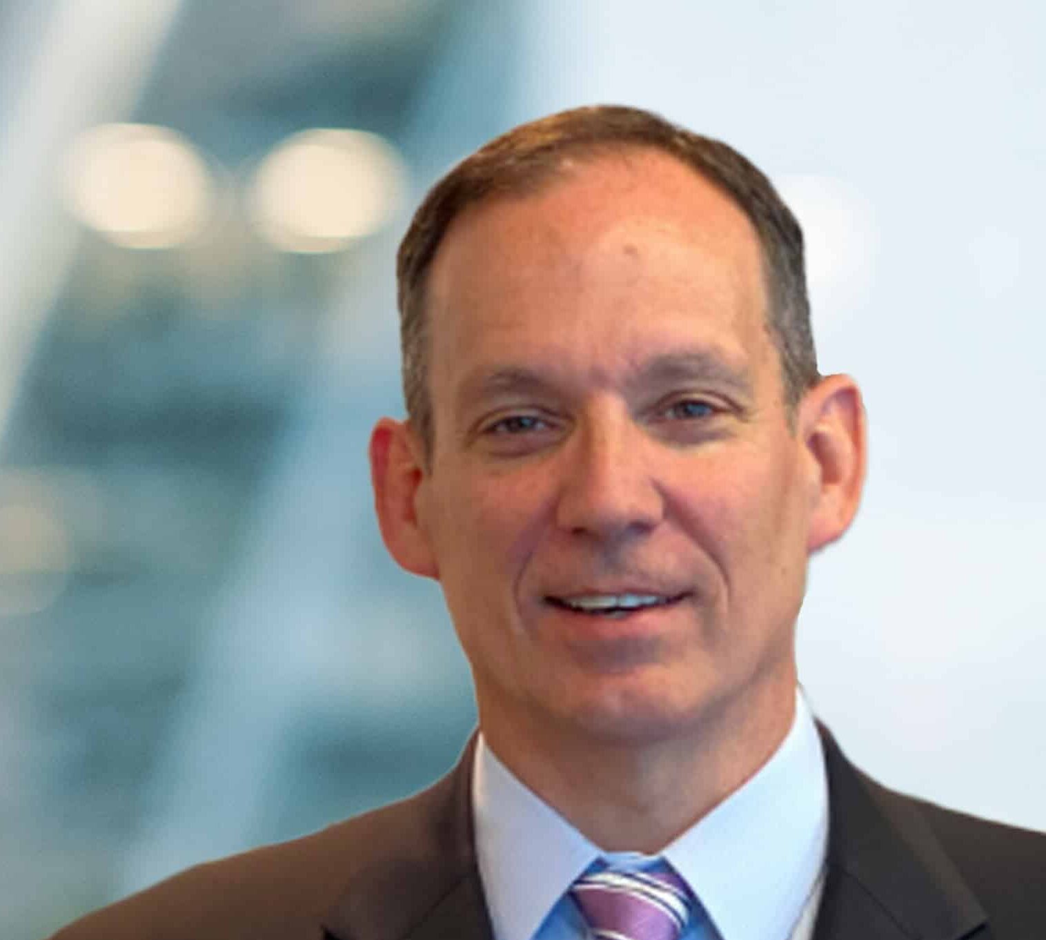 A man in a suit and tie smiles in front of a blurred, modern office background with soft lighting and glass architecture.