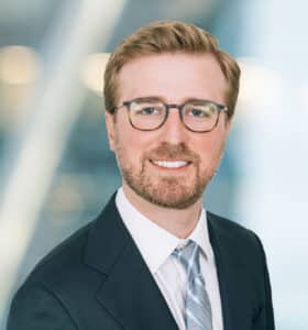 A man with light brown hair, a short beard, and glasses is wearing a dark suit, white shirt, and light-colored tie, smiling at the camera against a blurred, professional background.
