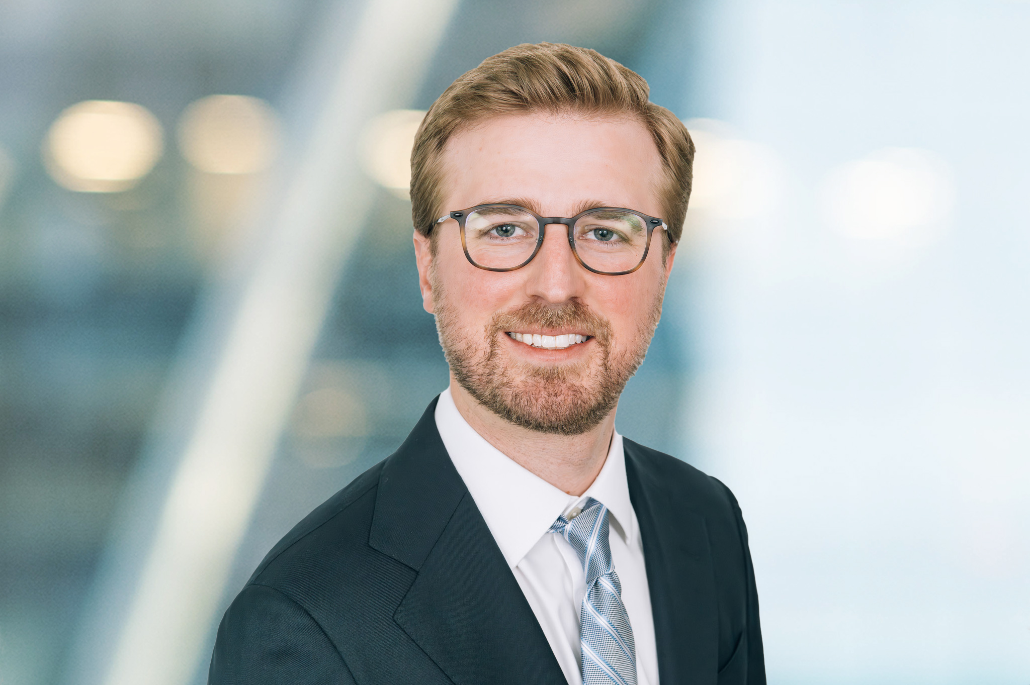 A man with light brown hair, a short beard, and glasses is wearing a dark suit, white shirt, and light-colored tie, smiling at the camera against a blurred, professional background.
