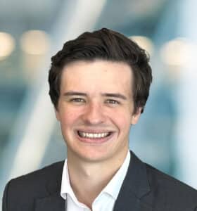 A young man with short brown hair, wearing a dark suit jacket and white shirt, smiles at the camera against a blurred, modern office background.