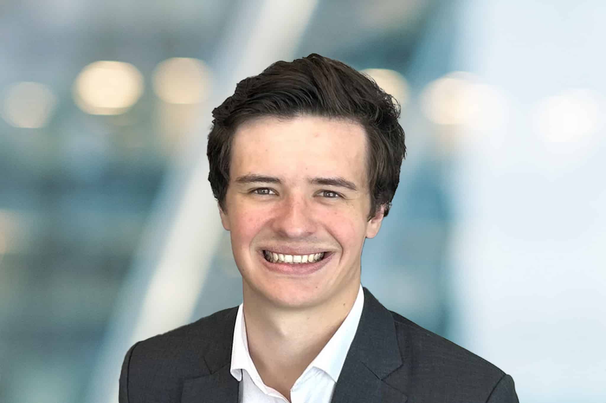 A young man with short brown hair, wearing a dark suit jacket and white shirt, smiles at the camera against a blurred, modern office background.