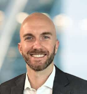 A smiling man with a bald head and beard, wearing a dark suit jacket and white shirt, stands in front of a blurred, modern indoor background with soft lighting.