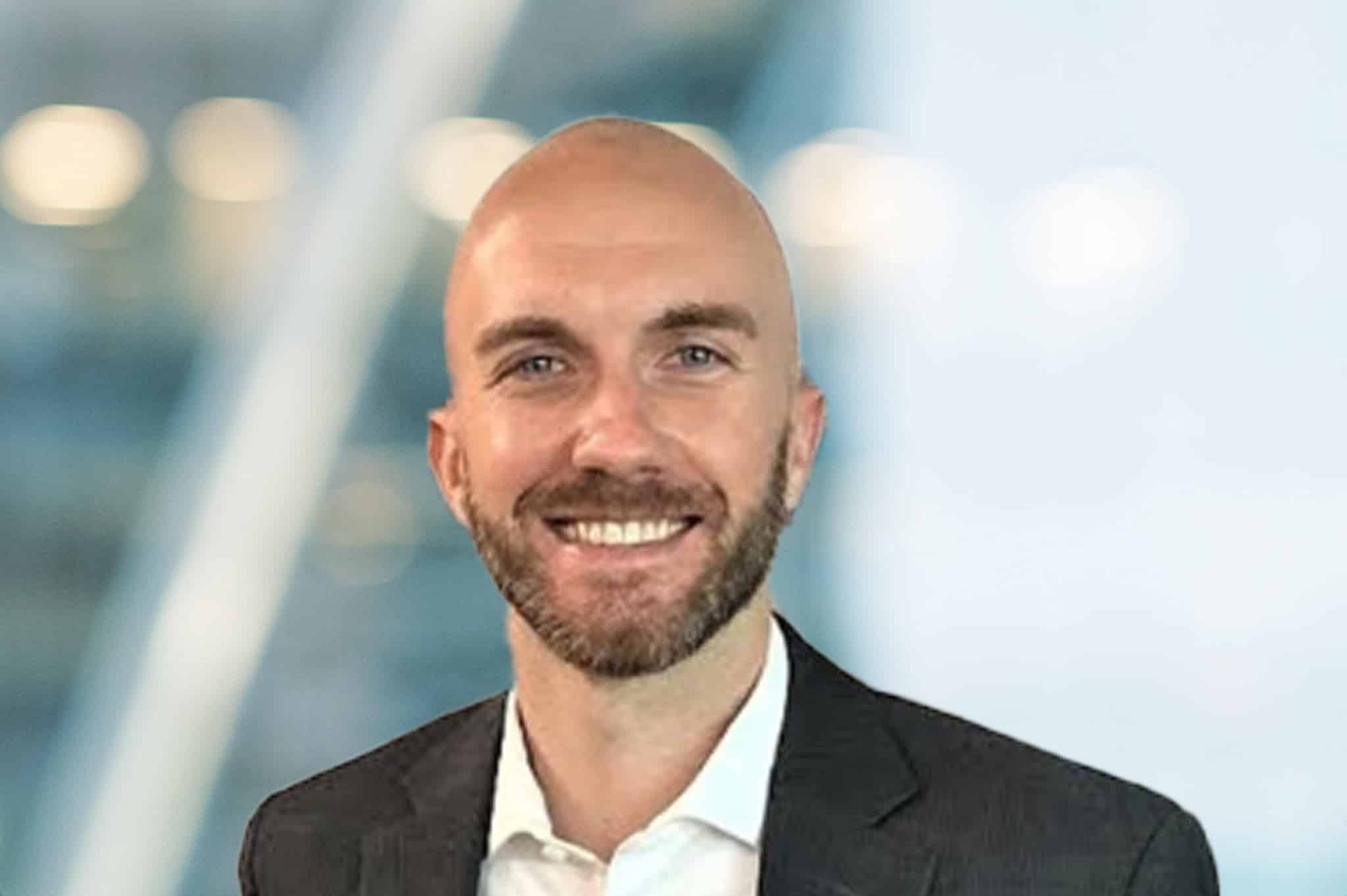 A smiling man with a bald head and beard, wearing a dark suit jacket and white shirt, stands in front of a blurred, modern indoor background with soft lighting.