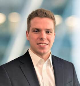 A young man with short light brown hair, wearing a black suit jacket and white dress shirt, smiles while standing in front of a blurred, modern office background.