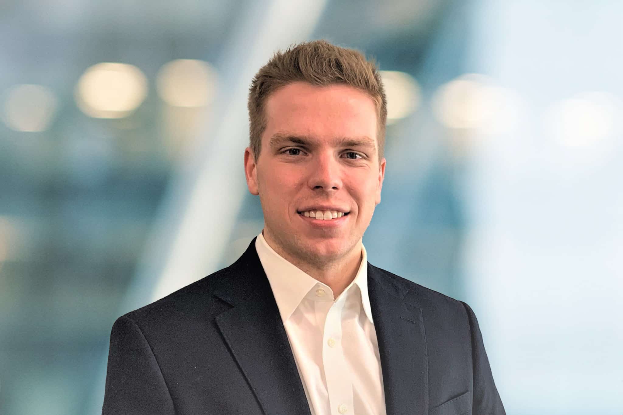 A young man with short light brown hair, wearing a black suit jacket and white dress shirt, smiles while standing in front of a blurred, modern office background.