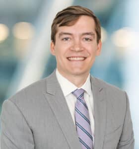 A man in a light gray suit, white shirt, and striped tie is smiling at the camera. The background is blurred with blue and white tones, giving a professional, modern office feel.