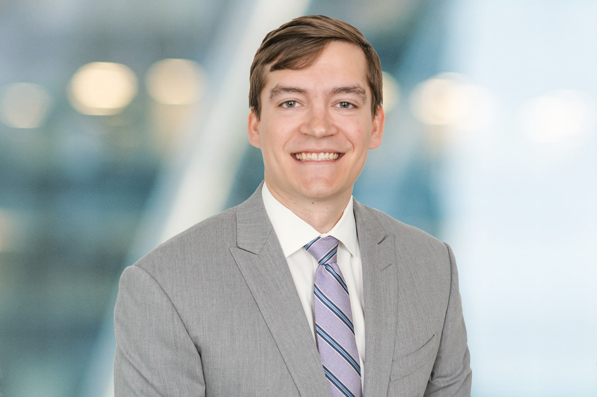 A man in a light gray suit, white shirt, and striped tie is smiling at the camera. The background is blurred with blue and white tones, giving a professional, modern office feel.