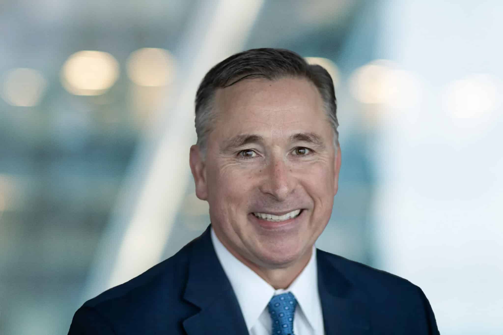 A man in a dark suit and blue tie smiles at the camera with a blurred background featuring soft lights and glass surfaces.