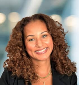 A woman with curly brown hair smiles at the camera, wearing a black blazer and layered gold necklaces, with a blurred background of lights and blue tones.