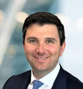 A man with short dark hair, wearing a dark suit, white shirt, and light blue tie, smiles at the camera. The background is blurred with soft blue and white tones.