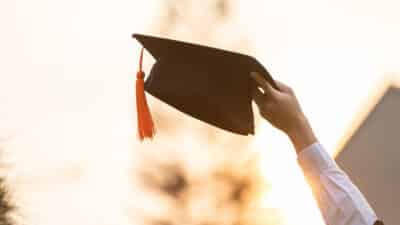 A person holds up a black graduation cap with an orange tassel against a bright, blurred outdoor background, symbolizing achievement and celebration.