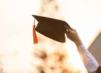 A person holds up a black graduation cap with an orange tassel against a bright, blurred outdoor background, symbolizing achievement and celebration.
