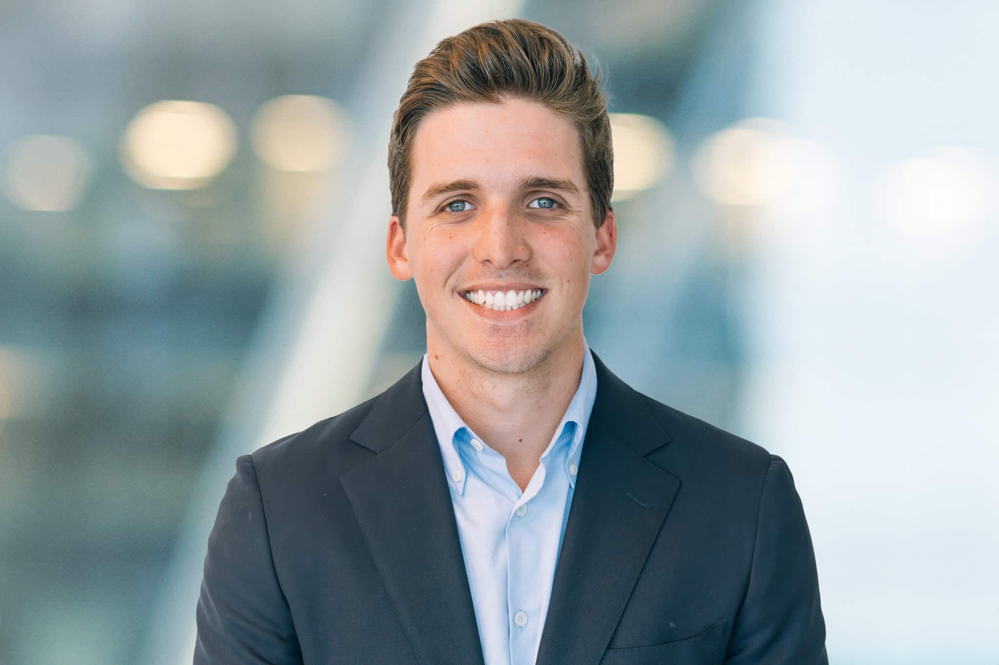A young man with short brown hair, wearing a dark suit jacket and light blue shirt, stands smiling in front of a blurred modern background with soft lights.
