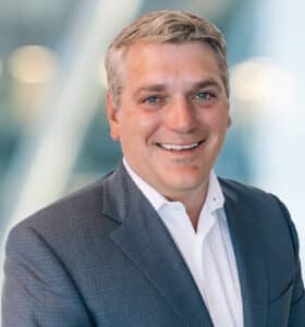 A man with short gray hair wearing a dark suit jacket and white dress shirt smiles at the camera against a blurred background with soft lights.
