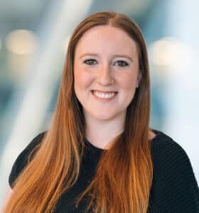 A woman with long red hair, wearing a black top, smiles at the camera. The background is softly blurred with blue and white tones, suggesting an indoor modern setting.