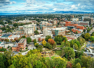 Aerial view of a cityscape with modern buildings, tree-lined streets, and a lush green park in the foreground under a partly cloudy sky.