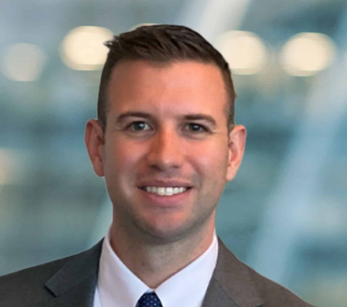 A man with short brown hair wearing a gray suit, white shirt, and dark tie smiles at the camera. The background is blurred with soft blue and white tones, suggesting an indoor office environment.