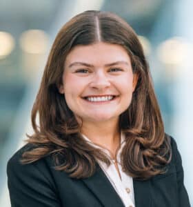 A woman with long brown hair, wearing a black blazer and white blouse, smiles at the camera. The background is blurred with soft lights and a modern indoor setting.