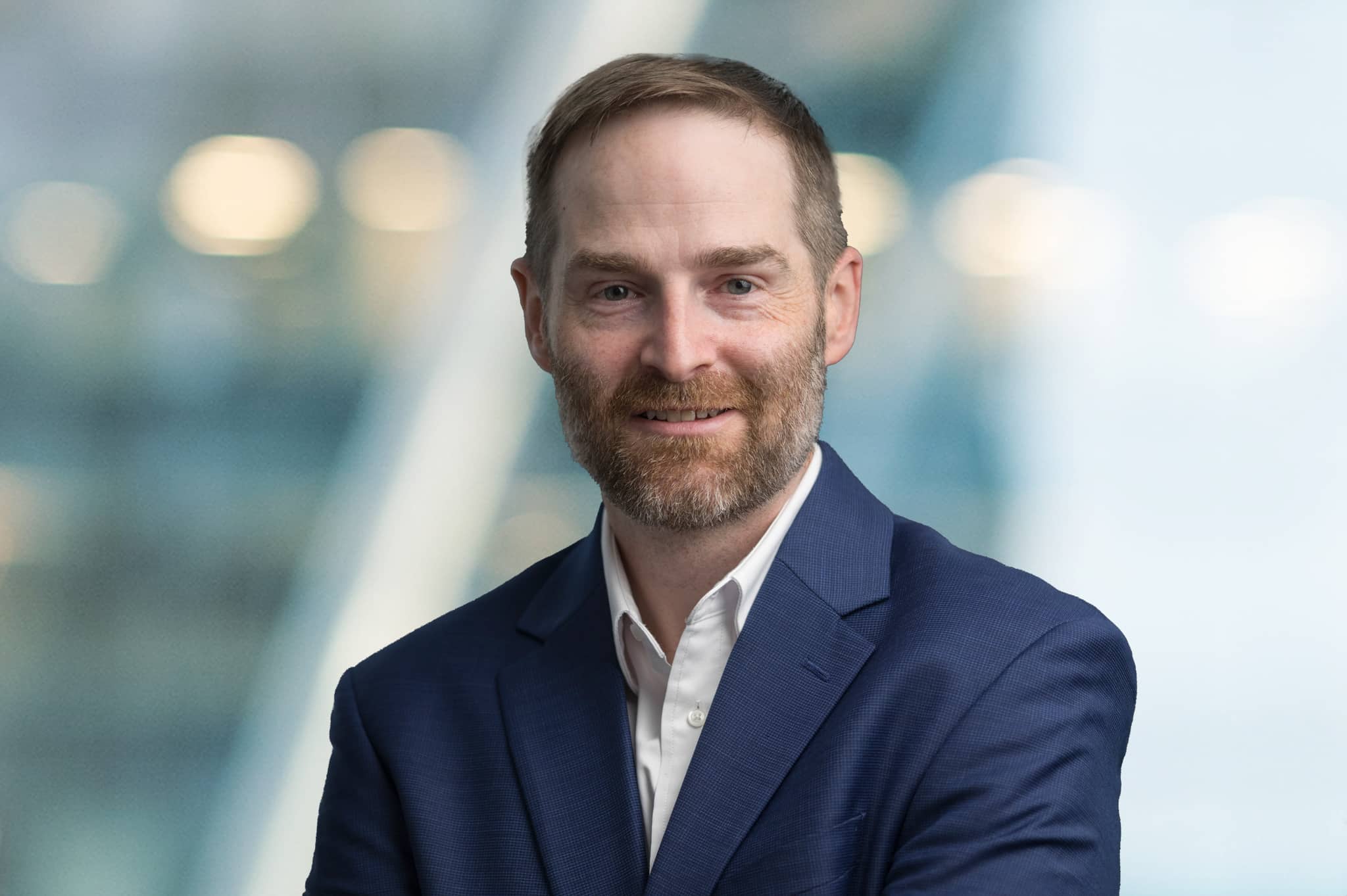 A man with short brown hair and a beard, wearing a navy blue suit and white shirt, smiles at the camera against a blurred background with soft lights.