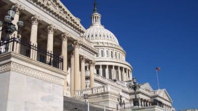 The United States Capitol building with its dome and columns, viewed from the steps, against a clear blue sky. The American flag is flying on a flagpole to the right.