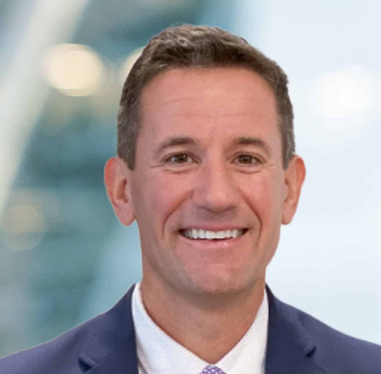 A man in a suit and tie smiles at the camera in front of a blurred, modern office background with soft lighting.