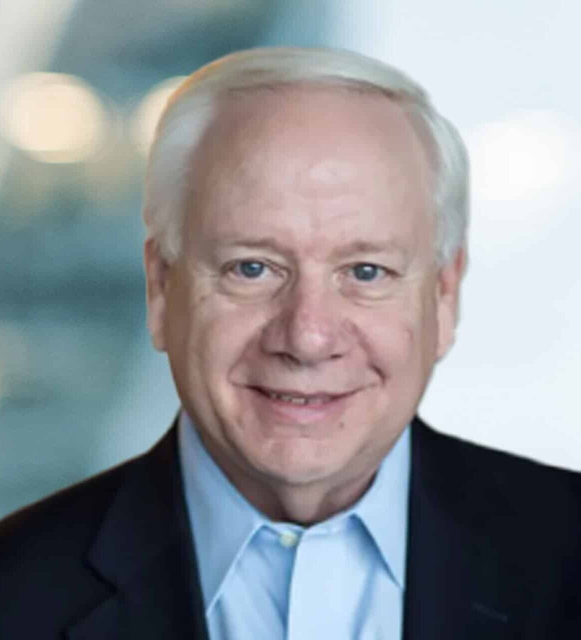 An older man with short white hair, wearing a dark suit jacket and light blue shirt, smiles at the camera against a blurred background with lights and glass panels.